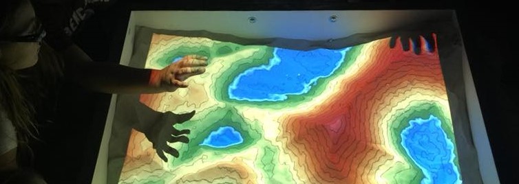 A close up photo of people interacting with an AR sandbox, showing color-coded topography